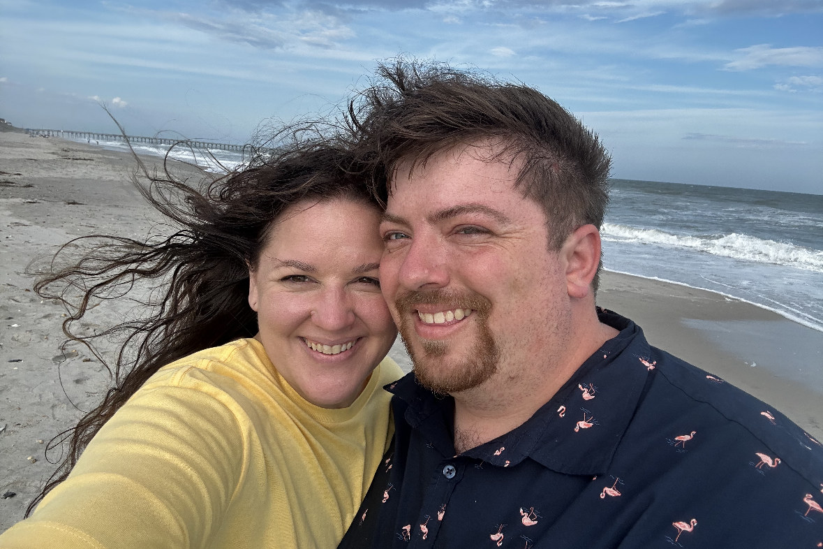 Kelly and Jessica cheek to cheek on a windy beach, the surf and a pier behind them.